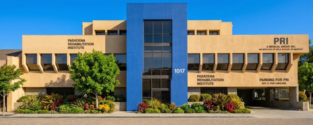 A symmetrically composed, direct frontal photograph of a modern medical facility under a vibrant, clear blue sky. Bright, natural daylight evenly illuminates the building, enhancing the rich, saturated colors of the prominent central blue panel and the surrounding warm beige stucco. The composition is clean and architectural, with a foreground of neatly laid pavers and lush, colorful garden beds featuring diverse plants, adding a sense of careful maintenance. Clear, sharp text is visible across the facade: large letters on the upper-left read "PASADENA REHABILITATION INSTITUTE"; the central address "1017" is prominent on the blue block; and on the upper-right, distinct text includes "PRI A MEDICAL GROUP, INC." and "AN AFFILIATE OF UCLA SCHOOL OF MEDICINE," along with other service and parking information. The overall impression is one of professional clarity and inviting care.