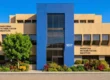 A symmetrically composed, direct frontal photograph of a modern medical facility under a vibrant, clear blue sky. Bright, natural daylight evenly illuminates the building, enhancing the rich, saturated colors of the prominent central blue panel and the surrounding warm beige stucco. The composition is clean and architectural, with a foreground of neatly laid pavers and lush, colorful garden beds featuring diverse plants, adding a sense of careful maintenance. Clear, sharp text is visible across the facade: large letters on the upper-left read "PASADENA REHABILITATION INSTITUTE"; the central address "1017" is prominent on the blue block; and on the upper-right, distinct text includes "PRI A MEDICAL GROUP, INC." and "AN AFFILIATE OF UCLA SCHOOL OF MEDICINE," along with other service and parking information. The overall impression is one of professional clarity and inviting care.