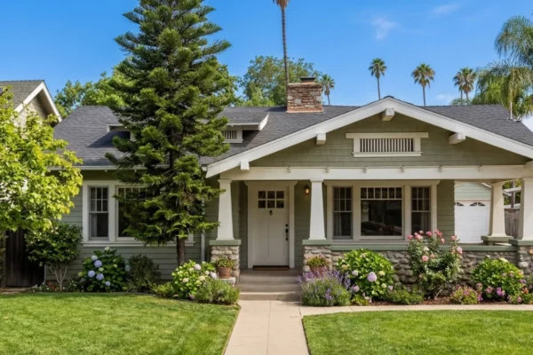 A wide, eye-level shot of a sage-green Craftsman bungalow bathed in bright, midday sunlight. The composition is balanced, featuring a symmetrical concrete path leading the eye toward a crisp white front door. High-contrast lighting emphasizes the textures of the stone-based pillars and wooden siding. Lush greenery and vibrant purple lavender frame the porch, while a towering, slender evergreen adds a vertical element against a clear, deep blue sky. The overall atmosphere is warm, serene, and suburban.