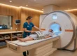 A wide, clean shot of a modern MRI suite with warm wood-paneled walls and recessed ceiling lights. A female technologist in teal scrubs is adjusting a coil over a patient lying on the table of a large white Siemens Magnetom Vida 3T machine. The branding "SIEMENS" and "MAGNETOM Vida 3T" is clearly visible on the front of the scanner. The composition is symmetrical and professional.