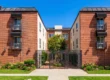 Bright, direct sunlight illuminates a symmetrical, three-story residential building facade under a flawless blue sky. The composition is perfectly balanced, featuring two identical red-brick wings with detailed dark iron balconies and awnings. These wings frame a central courtyard structure with a wrought-iron gate and walkway, creating a sense of balanced depth. Lush green trees, bushes, and flowering garden beds line the neatly paved sidewalk. The overall look is structured, well-kept, and brightly lit, emphasizing the textures of brick, metal, and foliage.