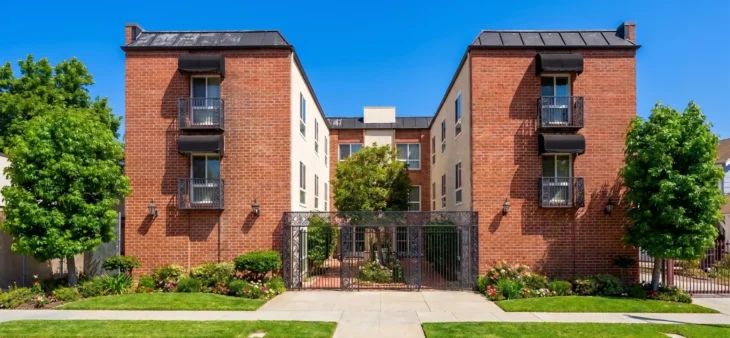 Bright, direct sunlight illuminates a symmetrical, three-story residential building facade under a flawless blue sky. The composition is perfectly balanced, featuring two identical red-brick wings with detailed dark iron balconies and awnings. These wings frame a central courtyard structure with a wrought-iron gate and walkway, creating a sense of balanced depth. Lush green trees, bushes, and flowering garden beds line the neatly paved sidewalk. The overall look is structured, well-kept, and brightly lit, emphasizing the textures of brick, metal, and foliage.
