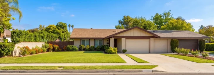 A perfectly centered, symmetrical wide-angle shot of a suburban home bathed in warm, bright daylight. The composition is flat and front-facing, emphasizing the house's clean beige walls and brown shingle roof against a vivid blue sky. Sharp details show the house number 172 above the entryway. The foreground features a vibrant, saturated green lawn and colorful flower beds, with soft, realistic shadows adding depth to the crisp architectural layout.