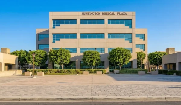 A symmetric, full-frontal photograph of a multi-tiered contemporary medical building facade with beige stucco walls and dark-framed reflective windows, illuminated by warm, late-afternoon golden sunlight. Dark text on the upper level reads 'HUNTINGTON MEDICAL PLAZA'. The building sits behind a wide, paved plaza featuring neatly spaced trees in square planters and modern lampposts. The background is a clear, deep blue sky. Long shadows extend across the foreground pavement.