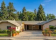 Bright clear daylight illuminates a frontal, centered wide-angle view of a cream-colored ranch-style facility. A concrete driveway leads under a central covered carport. Rows of terra-cotta pots with vibrant magenta and red flowering plants line the drive, flanked by chain-link fences and green lawns. The multi-winged building with a grey tile roof sits against a dense backdrop of mature pine, evergreen, and cypress trees under a clear blue sky. No text is visible on the structure.