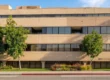 A front-on, horizontal photograph of a multi-story, sand-colored medical office building at 301 S Fair Oaks Ave, Pasadena, captured in bright natural daylight. The composition features a row of mature trees and landscaping along the green lawn in the foreground. Wide ribbon windows reflect the blue sky. A rounded glass corner with a tall palm tree is on the right. A leasing sign on the lawn displays legible text, including '301'.