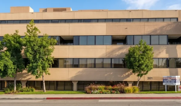 A front-on, horizontal photograph of a multi-story, sand-colored medical office building at 301 S Fair Oaks Ave, Pasadena, captured in bright natural daylight. The composition features a row of mature trees and landscaping along the green lawn in the foreground. Wide ribbon windows reflect the blue sky. A rounded glass corner with a tall palm tree is on the right. A leasing sign on the lawn displays legible text, including '301'.