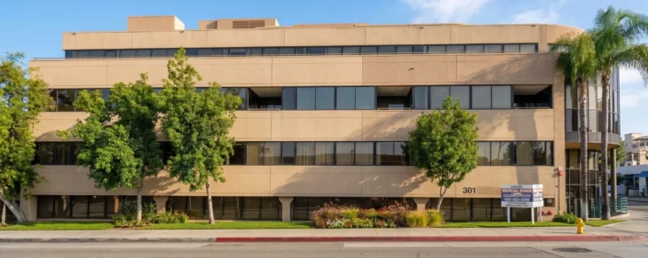A front-on, horizontal photograph of a multi-story, sand-colored medical office building at 301 S Fair Oaks Ave, Pasadena, captured in bright natural daylight. The composition features a row of mature trees and landscaping along the green lawn in the foreground. Wide ribbon windows reflect the blue sky. A rounded glass corner with a tall palm tree is on the right. A leasing sign on the lawn displays legible text, including '301'.