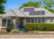 Single-story stucco home with a manicured hedge, bay window, and rooftop solar panels under bright blue California sky — address marker 2135 visible on the side wall, warm natural daylight, symmetrical composition with mature trees framing the entrance.