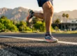 A detailed, low-angle photograph of a person's legs and feet running on an asphalt road marked with a white painted line. The scene is bathed in warm, golden hour sunlight, highlighting the musculature, skin texture, and the mesh of the grey and orange-detailed running shoes. The forward leg is bent, planting a foot on the ground, while the rear foot is lifted in mid-stride. In the softly blurred background, under rugged mountains and a green landscape with palm trees, a grand building with visible text 'ROSE BOWL' is set. The focus is sharp on the runner's legs, with a shallow depth of field for the background.