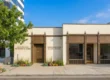 A wide, sun-drenched exterior photograph of the Tao of Wellness clinic in Pasadena, shot from across a clean sidewalk. The charming, cream-colored stucco building is framed by a mature green tree and low, textured desert plantings. Detailed, dark brown ornamental plasterwork trims the two storefront entrances and large arched windows, contrasting sharply against the light facade and clear blue sky. On the left section of the building, above a brown door, the text "ACUPUNCTURE NUTRITION & HERBS" is legible. On the right, above the main entrance window with decorative columns, the text "CHINESE MEDICINE QI GONG STUDIO" is displayed, with the address number "171" centered below.