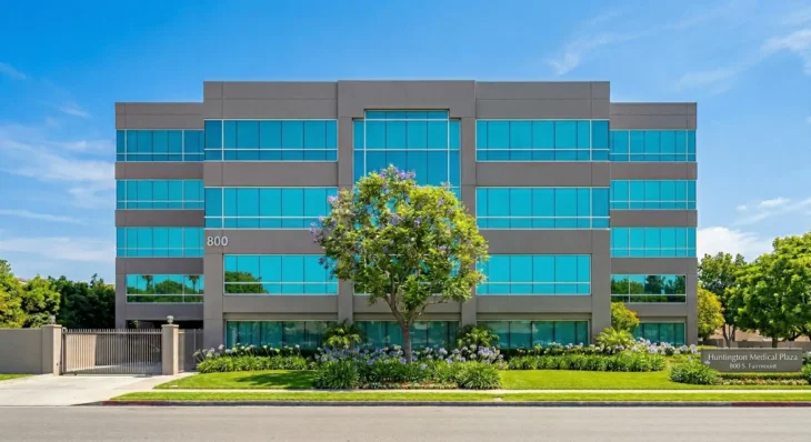 A symmetric, full frontal photograph of a modern multi-story medical office building under a bright blue sky with wispy clouds. The gray and taupe facade features rows of turquoise-tinted glass windows reflecting the clear daylight. A solitary tree, in purple bloom, is positioned precisely in the center foreground. A manicured lawn and garden bed with purple agapanthus flowers lead up to the building entrance. The building itself bears the large silver numbers "800" on the left side, on the second story. A ground-level, dark brown sign in the right foreground contains the gold-lettered text "Huntington Medical Plaza" above "800 S. Fairmount".