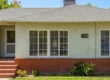 A centered, wide-angle exterior photograph of a single-story bungalow under bright, natural daylight, bathed in even, front-facing light. The balanced composition features a light-mint green stucco facade set upon a red brick foundation. Centered on the mint green wall, the text "1744" is painted in black characters. From the left, a path and red brick steps lead to a glass-paneled front door with a lantern. The center and right are occupied by windows with white grid patterns, one large and one smaller. Dense green trees fill the background under a blue sky, with varied low-lying plants in the foreground lawn.