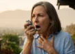 Close-up portrait composition with a shallow depth of field, contrasting a sharply focused middle-aged woman against a soft, heavily blurred background of smog-obscured mountains. The scene is illuminated by warm, diffused late-afternoon sunlight filtering through thick atmospheric haze. The woman is captured in a moment of acute physical distress, illustrating severe asthma symptoms; her mouth is open as she clutches her chest with one hand. On the other hand, she holds a blue L-shaped asthma medication inhaler—featuring small, blurred white printed text on its metallic canister—directly to her mouth, visually representing an asthma attack triggered by air pollution