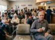 A deeply layered, wide-angle interior photograph of a crowded urgent care waiting room in Pasadena. Natural daylight from a wall of windows on the left mixes with warm overhead artificial lighting, illuminating varied areas of the room. In the immediate foreground, an elderly man in a grey-and-blue plaid shirt is seated, holding a paper ticket clearly reading “#58” and looking directly at the camera. Rows of diverse patients of varying ages fill the midground, seated in rows of grey chairs, some using phones, including a woman holding a baby. A dense queue of standing people on the right leads back to a brightly lit reception desk with a waiting line. Above the counter, prominent hanging signs include 'CHECK-IN' in white letters on blue, and a large red sign with 'WAITING TIMES: >3 HRS' in large white text. A 'PASADENA URGENT CARE' sign is on a wall-mounted poster and the red header. Wall posters include one with a photo of a doctor. The shot creates a profound sense of scale and human congestion.