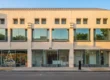 A perfectly centered, straight-on frontal composition of a two-story commercial building bathed in warm, bright natural daylight. The lighting is vibrant and clear, casting soft, realistic shadows against the beige facade under a clean blue sky. The shot features high contrast and professional sharpness. Text visible on the building includes a vertical "FOR LEASE" sign on the upper left side, and a red-and-white "AVAILABLE" sign with a phone number on the far right structural pillar.