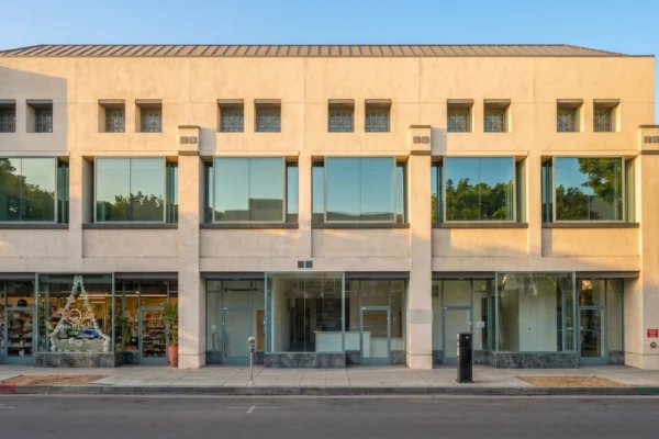 A perfectly centered, straight-on frontal composition of a two-story commercial building bathed in warm, bright natural daylight. The lighting is vibrant and clear, casting soft, realistic shadows against the beige facade under a clean blue sky. The shot features high contrast and professional sharpness. Text visible on the building includes a vertical "FOR LEASE" sign on the upper left side, and a red-and-white "AVAILABLE" sign with a phone number on the far right structural pillar.