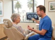 A softly lit photograph bathed in warm, natural light, using a medium side-angle composition to highlight a dental consultation. A dentist in scrubs is positioned next to a senior patient seated in a dental chair. The visual focus is on a small, detailed titanium and white dental implant model held in the dentist's hand, which both individuals are looking at attentively. In the softly blurred background, a glowing monitor displays a black-and-white panoramic jaw X-ray. The scene is modern and inviting, with no visible text.
