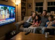 Cinematic low-key lighting with warm ambient room lamps contrasting against the cool, blue illumination cast by a large television screen onto a tense family of four. The medium-shot composition balances the brightly lit screen on the left with the huddled family seated on a sofa on the right. The TV displays a news broadcast featuring workers in hazmat suits and a chyron with the exact text: "NEWS LIVE ARDBREAKS", "EBOLA OUTBREAK SPREADS: GLOBAL ALERT", and "HEALTH EMERGENCY DECLARED IN SEVERAL COUNTRIES".