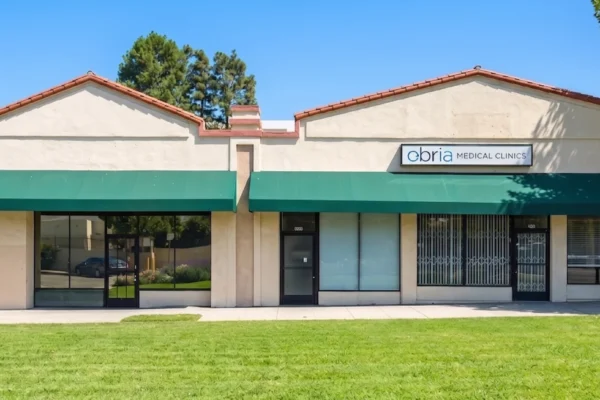 A sharp, frontal photograph of a symmetrical single-story commercial building under a clear, bright blue sky with direct, crisp sunlight. The building features two peaks with terracotta tile trim, cream stucco walls, and continuous forest green awnings stretching across the facade. The foreground is filled by a wide, vibrant green lawn and a concrete sidewalk. Large glass windows and doors stretch across the width, and on the right gable, a prominent white rectangular sign displays the text "obria MEDICAL CLINICS" in blue and black letters.