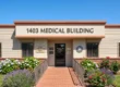 A perfectly centered, symmetrical architectural composition illuminated by bright, warm daylight under a clear, vibrant blue sky. A textured red brick pathway, bordered by tan metal handrails and lush green lawns with colorful flowering bushes, leads the eye directly to the central entrance of a single-story facade. Prominent dark brown block letters spelling "1403 MEDICAL BUILDING" are mounted across the upper exterior above the shaded glass double-door entrance, which displays smaller, less legible informational plaques. The crisp lighting creates soft, realistic shadows, enhancing the rich color contrast and inviting atmosphere of the scene.
