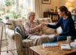 A detailed, wide-angle photograph capturing a tender moment in a cozy, lived-in living room. Warm, soft natural light streams in from a large garden-facing window on the left, illuminating a metal walker standing beside the window frame. An older woman with grey hair, glasses, and a floral top is seated comfortably on a large patterned sofa, covered by a blanket, smiling as she accepts a glass of water from a younger woman with dark hair and a blue sweater, who is kneeling at her side and smiling. The younger woman's kneeling posture and the older woman's seated position create a dynamic central focus of care. The foreground on a rustic wooden coffee table shows a light blue weekly pill organizer, a tissue box, a mug on a coaster, and a book titled "Recovery Journey." The background features extensive bookshelves packed with books and personal items, with lamps emitting a warm, inviting yellow glow, creating a cared-for and comfortable domestic scene.