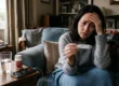 A shallow depth of field composition illuminated by soft, dim, diffused natural light originating from the left. The primary focal point is a digital thermometer held in the center foreground. Directly behind it, a woman's face is softly lit with gentle shadows on her right side, her hand resting against her forehead. The lower-left foreground features a wooden side table holding a glass of water, a smartphone with a dark screen displaying "16:04", and a medicine bottle bearing the text "TYLENOL". The background consists of a gently blurred living room with a textured blue couch and framed pictures.