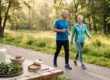 Soft, warm morning sunlight filters through lush green trees, creating a bright and encouraging atmosphere. The composition features a sharply focused foreground arrangement of a tall glass of milk, creamy yogurt, vibrant leafy greens, and a tin of salmon resting on a wooden surface. In the softly blurred background, an active older couple in athletic wear walks briskly from left to right, smiling warmly. There is no text present in the image.