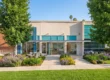 Bright, warm daylight illuminates the textured beige stucco and brick facade of a perfectly centered, modern commercial building. The composition is balanced and symmetrical, framed by two large mature deciduous trees on either side of a clean stone walkway. A well-maintained lawn and lush front garden with mixed flower beds containing purple lavender, yellow, and blue blooms lead to a glass-front entrance. The facade features distinct turquoise accent tiling and tinted windows. On the far left brick panel, the number '1595' is clearly visible. Centered above the main entrance doors, the text 'CHAPcare HEALTH CENTER' is present with a stylized cross logo.