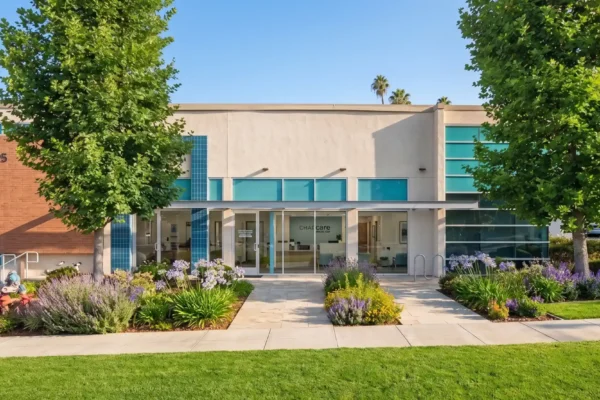 Bright, warm daylight illuminates the textured beige stucco and brick facade of a perfectly centered, modern commercial building. The composition is balanced and symmetrical, framed by two large mature deciduous trees on either side of a clean stone walkway. A well-maintained lawn and lush front garden with mixed flower beds containing purple lavender, yellow, and blue blooms lead to a glass-front entrance. The facade features distinct turquoise accent tiling and tinted windows. On the far left brick panel, the number '1595' is clearly visible. Centered above the main entrance doors, the text 'CHAPcare HEALTH CENTER' is present with a stylized cross logo.