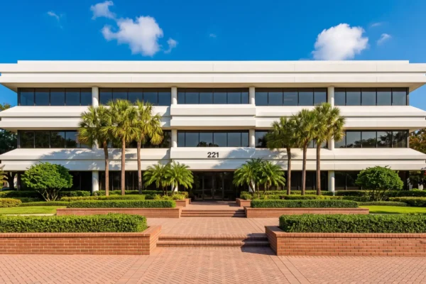 A symmetrical, head-on photograph under a clear blue sky with light clouds, showcasing a modern office building with multiple horizontal levels of alternating white stucco bands and dark ribbon windows. The building is framed by several tall palm trees. The foreground features tiered, well-manicured evergreen hedges and brick-paved walkways leading to a central glass entrance. The number "221" is clearly displayed in black above the entryway. The scene is brightly lit with even daylight.
