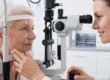 Soft, bright, even illumination fills a clean professional medical office. A complex grey and black slit-lamp machine is centered in the profile composition, flanked by an elderly woman with short white hair (left) and a younger female doctor with brown hair (right). Both are in profile, with the machine as the point of converge for their gaze and hands. The background is a gently blurred white space with visible cabinet handles and a distant medical chart containing small, illegible black text. The doctor's hand is on a machine control knob and also supporting the patient's chin rest.