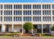 A perfectly centered, flat frontal architectural composition of a multi-story white building featuring a symmetrical grid of recessed rectangular windows. The scene is illuminated by bright, warm, natural sunlight that creates crisp, realistic shadows within the deep window bays, enhancing the building's geometric texture. The high-contrast, vibrant lighting is complemented by a clear, vivid blue sky at the top edge and lush, highly saturated green foliage framing the ground-level entrance. Faint text and typographic layouts are visible on the informational posters displayed behind the expansive, dark glass windows of the central ground-floor lobby.