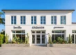 A perfectly centered, front-on architectural photograph of a sleek, two-story white stucco building under a clear blue sky. Bright daylight illuminates the symmetrical facade, defining columns and window frames. Below the rows of identical second-story windows, the distinct dark, modern text "breilla", "EREWHON", "prenuvo", and "tia" marks the storefronts. Ground-level planters filled with mature cacti like prickly pears and organ pipes line the concrete walk. A tall palm tree stands on the far left, balanced by lush tree cover on the far right. The lines are clean, and the overall composition is one of structured symmetry.
