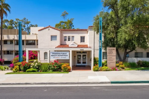 A wide, bright exterior photograph captures the Pasadena Community Center under a clear blue sky. The composition is a direct frontal view from a paved courtyard. In the foreground, a precise geometric pattern of stone pavers leads to a perfectly manicured lawn and colorful flower beds containing purple, pink, and white flowers. The central building, a two-story beige-painted structure with a Spanish-style red tile porch, is framed symmetrically by two modern, bright teal-blue vertical columns. Bright, direct sunlight from the right illuminates the stucco facade, creating sharp, soft shadows and making the colors vibrant. A cluster of very tall palm trees towers on the left, contrasting with a large, full, leafy oak-like tree on the right. A single ornate, vintage-style lamp post stands to the right of the sign.