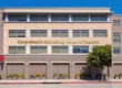 A wide-angle, horizontal photo under a clear blue sky, capturing the modern multi-story facade of a medical office building. Bright daylight casts clean shadows, emphasizing the grid of windows, light tan brick panels, and darker grey brick base. Prominent gold dimensional letters are centered on the upper brick section, spelling out: 'Comprehensive Dermatology Center of Pasadena'. Below, a row of uniform brick planters with low-lying flowers and shrubs lines the sidewalk. To the far left, a small, square red sign is visible. A leafy tree stands to the right, adding organic texture to the architectural lines.
