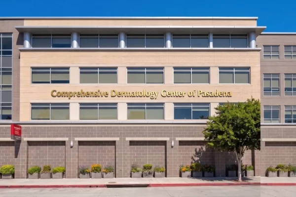 A wide-angle, horizontal photo under a clear blue sky, capturing the modern multi-story facade of a medical office building. Bright daylight casts clean shadows, emphasizing the grid of windows, light tan brick panels, and darker grey brick base. Prominent gold dimensional letters are centered on the upper brick section, spelling out: 'Comprehensive Dermatology Center of Pasadena'. Below, a row of uniform brick planters with low-lying flowers and shrubs lines the sidewalk. To the far left, a small, square red sign is visible. A leafy tree stands to the right, adding organic texture to the architectural lines.
