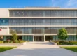A perfectly centered, straight-on architectural composition of a modern white and glass building captured under bright, vibrant daylight. Warm, natural sunlight casts soft, realistic shadows across the wide paved walkway and the flanking lush, saturated green landscaping. The lighting is crisp and high-contrast against a clear blue sky, giving it the polished look of professional real estate photography. On the lower left section of the facade, the text "Keck Medicine of USC" stands out clearly in bold black and red lettering.