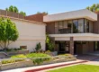 Bright daylight exterior photograph capturing a two-story commercial building with contrasting textures of beige stucco and red-brown brick. The eye-level composition features landscaped concrete planters with lush green shrubs and a small tree in the foreground. A paved parking area with a red-painted curb leads towards the entrance, casting strong sunlight and distinct shadows. Mature green trees are visible against a clear blue sky in the background.