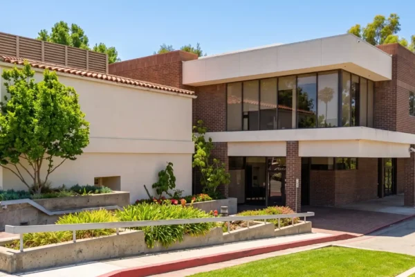 Bright daylight exterior photograph capturing a two-story commercial building with contrasting textures of beige stucco and red-brown brick. The eye-level composition features landscaped concrete planters with lush green shrubs and a small tree in the foreground. A paved parking area with a red-painted curb leads towards the entrance, casting strong sunlight and distinct shadows. Mature green trees are visible against a clear blue sky in the background.