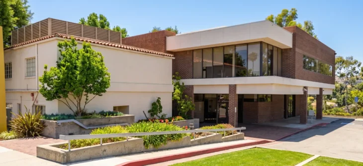Bright daylight exterior photograph capturing a two-story commercial building with contrasting textures of beige stucco and red-brown brick. The eye-level composition features landscaped concrete planters with lush green shrubs and a small tree in the foreground. A paved parking area with a red-painted curb leads towards the entrance, casting strong sunlight and distinct shadows. Mature green trees are visible against a clear blue sky in the background.