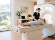 A wide, photorealistic composition bathed in soft, natural daylight streaming from a large window on the left, creating a warm, airy, and uplifting atmosphere. The scene features a shallow depth of field with vibrant, true-to-life colors. On the right, a pristine white MRI scanner with smooth, unbranded, and text-free surfaces anchors the frame. In the center, bathed in the gentle light, a smiling technologist in dark blue scrubs interacts with a patient lying on the extended scanner bed, set against a clean, minimalist background.