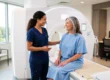 A brightly lit, sun-drenched composition featuring a large, modern white medical scanner as the central backdrop. Soft, warm natural light streams from large windows on the left side, evenly illuminating the two figures in the foreground. The shallow depth of field softly blurs the background office elements and seating area, drawing focus to the clean, inviting center of the frame. There is no visible text on the equipment or garments.