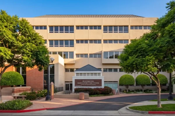 A perfectly centered, front-facing architectural photograph bathed in bright, warm natural sunlight under a clear blue sky. The symmetrical composition features vibrant, lush green trees flanking a multi-story beige building. In the immediate foreground, a crisp, dark-colored monument sign displays the text 'HUNTINGTON AMBULATORY CARE CENTER' in clean white typography. The image boasts high contrast, soft realistic shadows, and a polished, professional real estate aesthetic.