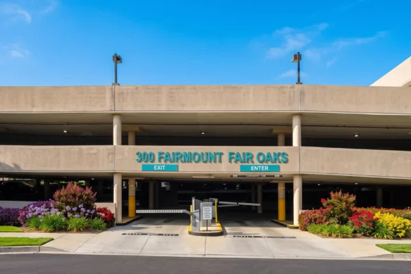 A brightly lit, symmetrical photograph of a multi-story concrete parking structure under a clear blue sky. The composition is frontal, featuring a central entrance area with automated barrier gates, flanked by vibrant, manicured flower beds with purple, red, and yellow blooms, and lush green trees on both sides. A teal-colored text sign centered on the facade reads '300 FAIRMOUNT FAIR OAKS', with smaller 'EXIT' and 'ENTER' signs below.